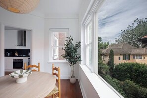 A room with a dining table, chairs, a potted plant, and a view of a house through a window. - Winchester Stylish Art Deco Apartment in Edgecliff (Edgecliff)
