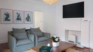 Een woonkamer met twee grijze banken, een houten salontafel, een boek, een witte vaas met bloemen, een flatscreen-televisie en een wit bakstenen haardvuur.