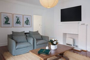 Ein Wohnzimmer mit zwei grauen Sofas, einem Holztisch, einem Buch, einer weißen Vase mit Blumen, einem Flachbildfernseher und einem Kamin aus weißen Ziegelsteinen.