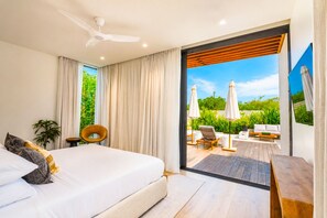 A bedroom with a large bed, a ceiling fan, and a view of an outdoor patio with seating and umbrellas.