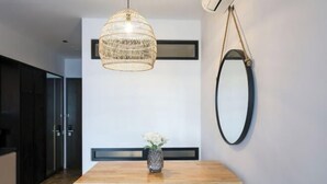 Uma área de refeições com mesa de madeira, duas cadeiras, luminária de vime pendente, espelho fixado na parede e um vaso com flores.