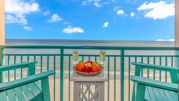 Een balkon met uitzicht op het strand, een tafel met een fruitmand en twee glazen wijn.