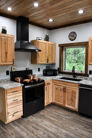 A kitchen with wooden cabinets, a black stove, and a window with a view of greenery. - Midnight Sun Retreat. 2 Minutes from Airport! (Fairbanks)