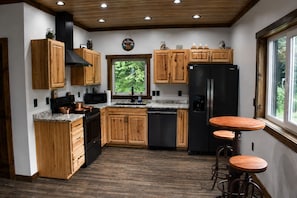 Una cocina con armarios de madera, un refrigerador negro, utensilios de cocina de cobre y una ventana con vista a follaje.