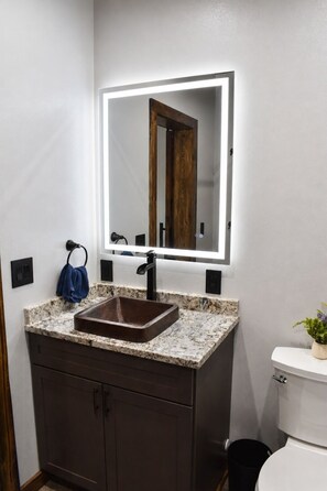 A bathroom with a marble countertop, a vessel sink, a wall-mounted mirror with lighting, a dark wooden cabinet, and a white toilet. - Midnight Sun Retreat. 2 Minutes from Airport! (Fairbanks)
