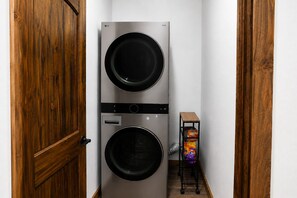 A laundry room with two stacked washing machines, a small shelf with detergent, and a wooden door. - Midnight Sun Retreat. 2 Minutes from Airport! (Fairbanks)