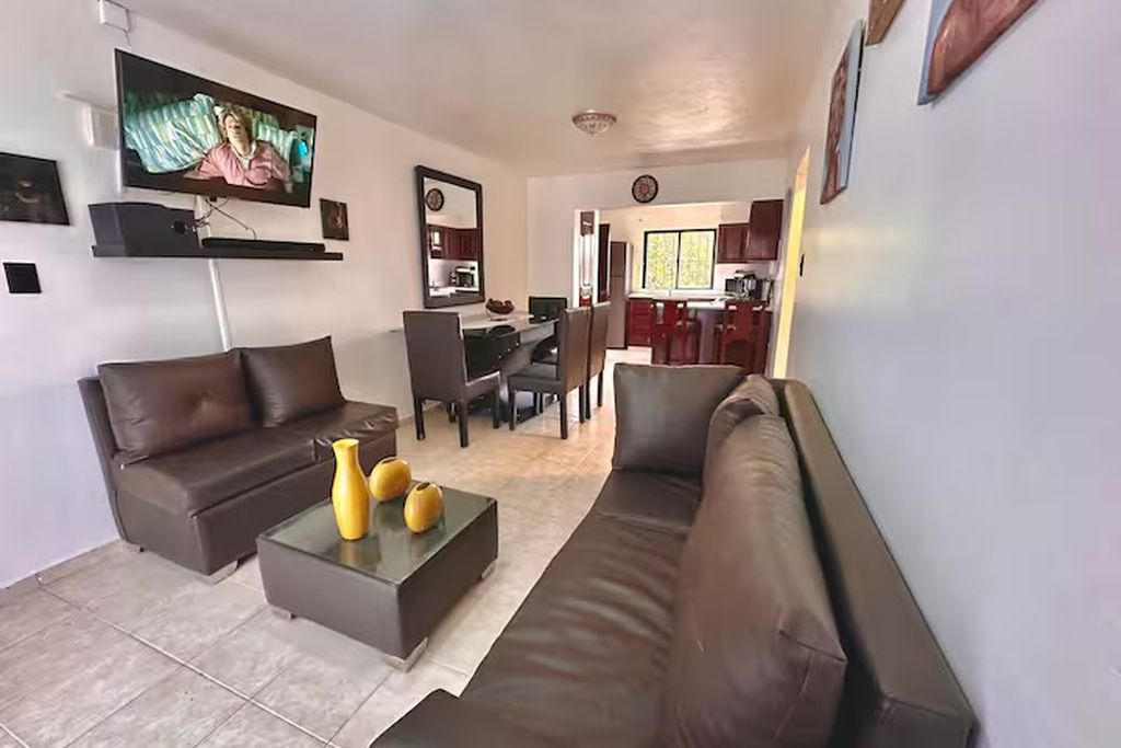 A modern living room with a flat-screen TV, two sofas, a coffee table, and a dining area in the background.