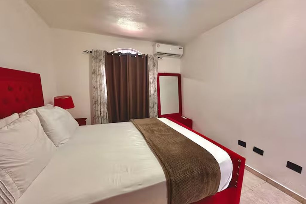 A hotel room with a bed, a red headboard, a bedside table with a lamp, a window with curtains, and an air conditioning unit.