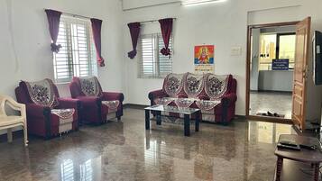 Amplia sala de estar con dos sofás rojos, una mesa de centro y una silla blanca. Hay ventanas con cortinas, un cartel en la pared y una puerta que da al exterior.