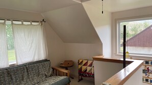 Una habitación con techo inclinado, un banco de madera, una mesita pequeña, un colcha de colores y una ventana que da a una vista de un edificio.