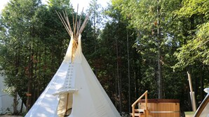 A white teepee with a ladder, wooden steps, and a hot tub in a forest setting.