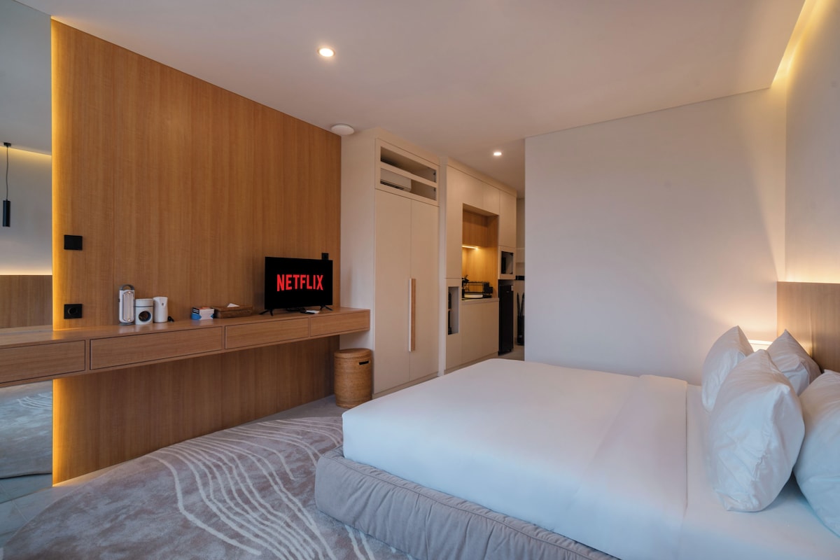 Ein modernes Hotelzimmer mit einem großen Bett, einem Holztisch, einem Fernseher mit 'NETFLIX' und einer Küchenzeile.