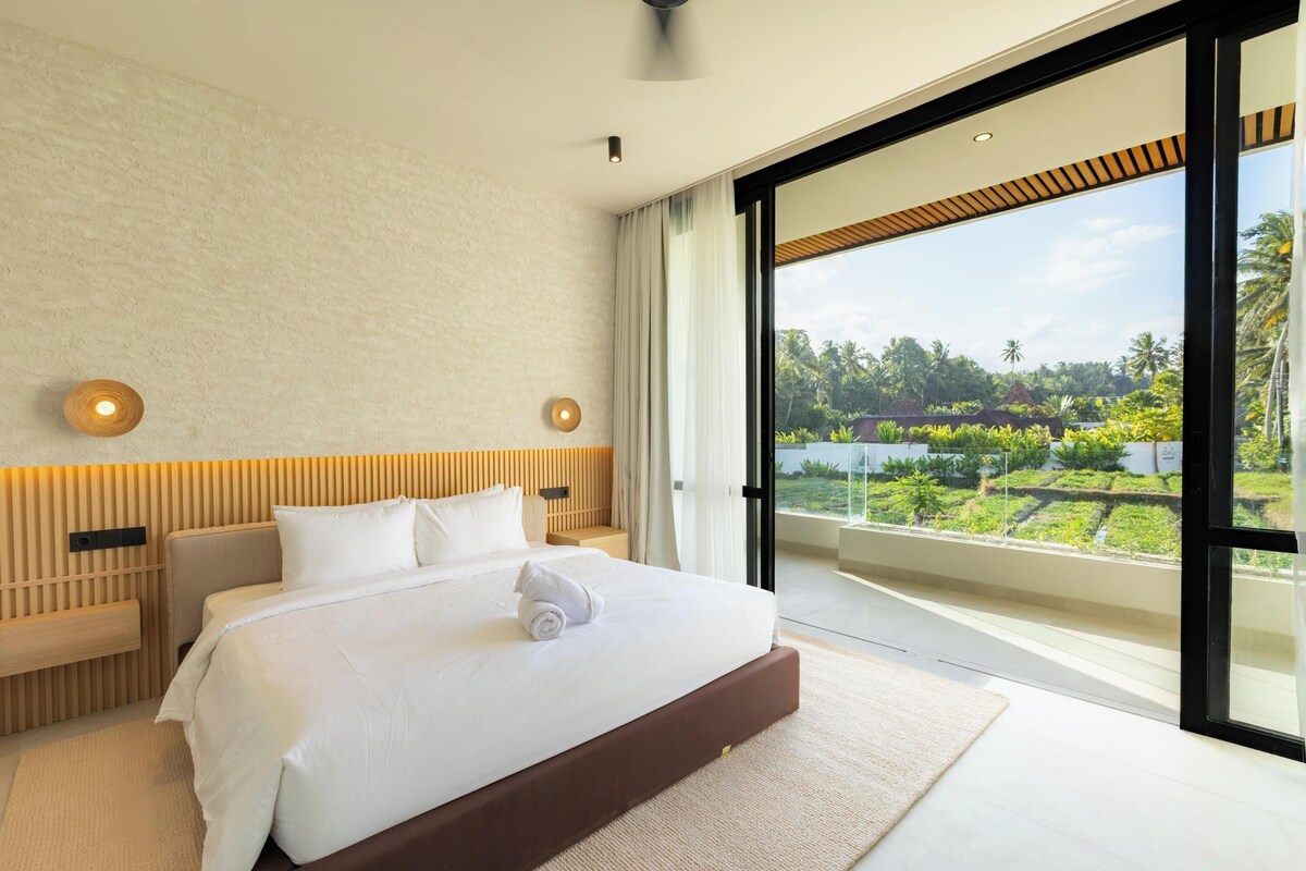 Quarto moderno com uma cama grande, cabeceira de madeira, mesa de cabeceira e vista para um jardim exuberante através de uma porta de vidro.