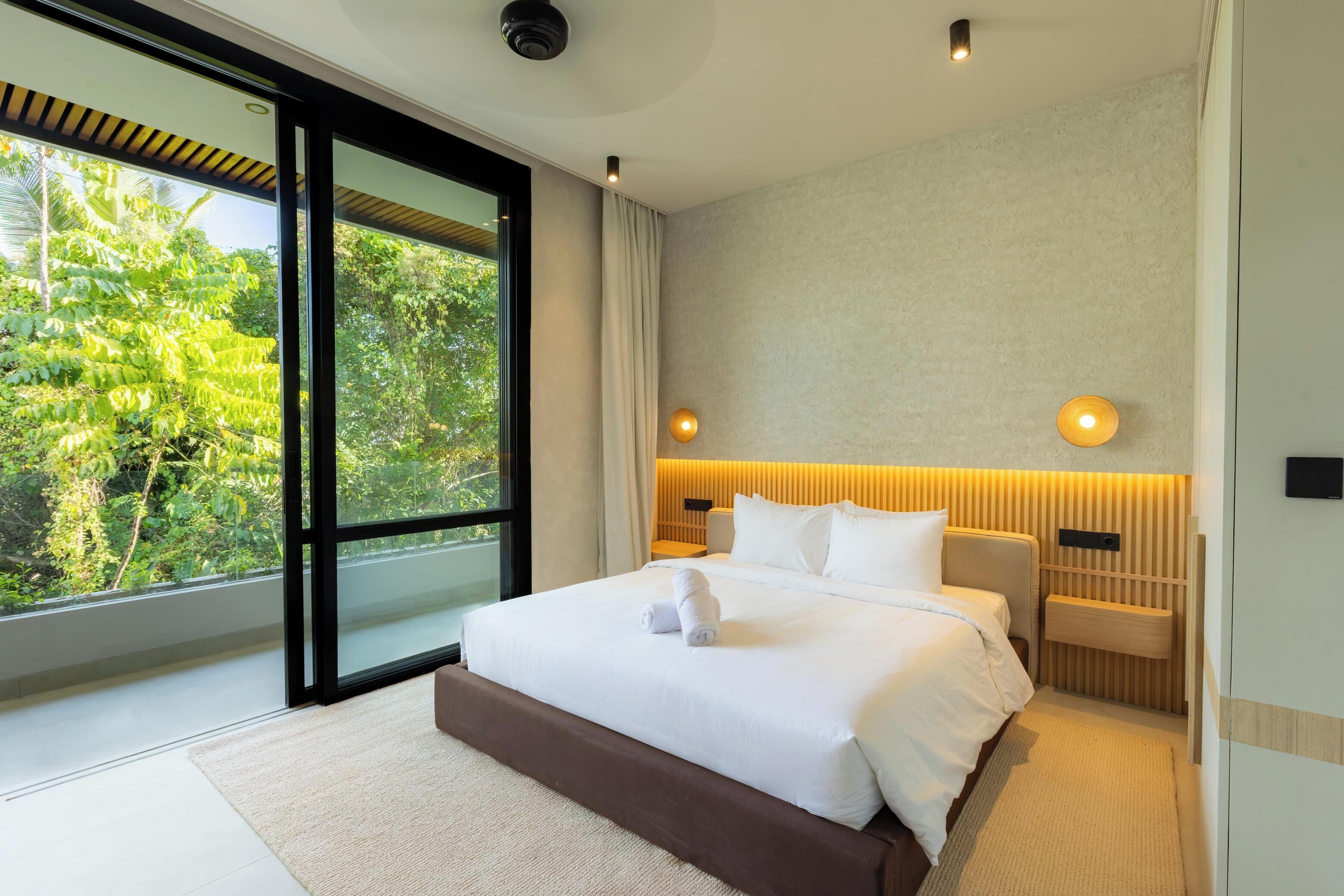 Una habitación de hotel moderna con una cama grande, vistas a zonas verdes a través de ventanales del suelo al techo y un cabecero de madera.