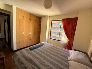 Ein Schlafzimmer mit großem Kleiderschrank, einem Bett mit gestreifter Bettwäsche, einem Fenster mit roten Vorhängen und Blick auf Gebäude.
