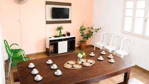 Quarto com mesa de madeira posta para o chá, TV fixada na parede, um armário e um vaso com planta.