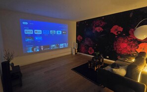 Ein Wohnzimmer mit einem großen Bildschirm, der eine Medien-App-Oberfläche zeigt, einer Wand mit dunklem Blumenmuster, einem Sofa und einem kleinen Tisch mit einer Lampe.