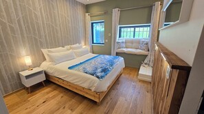 Ein Schlafzimmer mit einem hölzernen Bett, weißer Bettwäsche, einem Nachttisch, einem Fenster mit Vorhängen und Blick ins Freie.