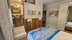 A bedroom with a bed, a wooden wardrobe, a bathroom with a sink and mirror, and a wall with a floral artwork.
