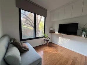Una sala de estar moderna con un sofá gris, un televisor montado en la pared y un ventanal con vista a árboles.