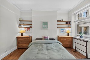 A bedroom with a bed, two nightstands, a wall-mounted shelf, a window with a view, and a 'JUST BE' sign. - Stylish Two Bedroom Apartment (London)