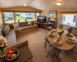Un salon chaleureux avec un canapé, un téléviseur, une cheminée, une table à manger et une vue sur une maison, visible à travers de grandes fenêtres.