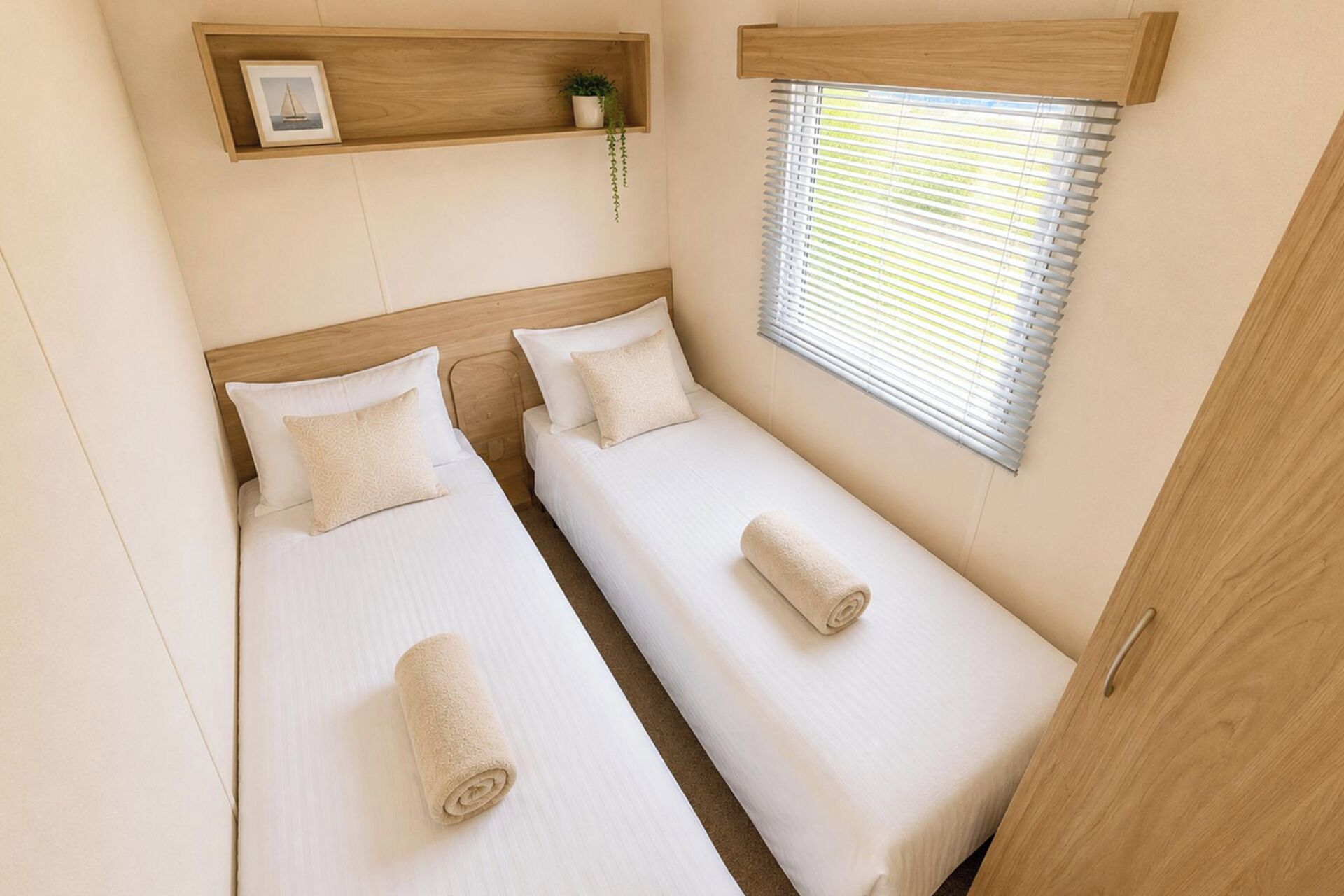 Een kleine, nette kamer met twee enkele bedden, een houten hoofdbord, een raam met zonwering en een plankje met een plant.