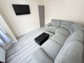 Ein Wohnzimmer mit Ecksofa, flachbildfernseher an der Wand und einer Tür.
