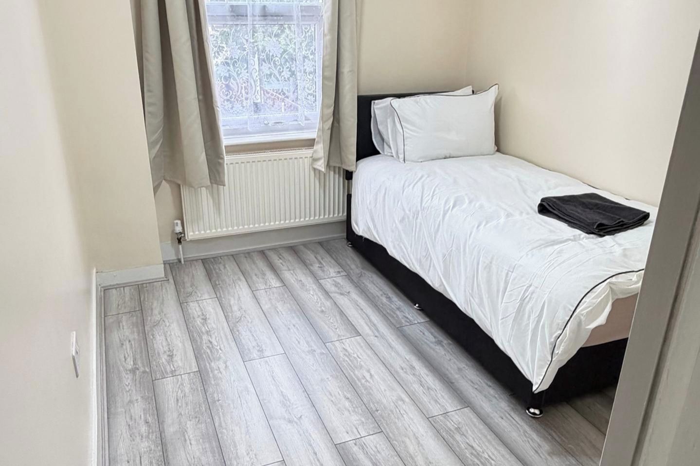Een kleine kamer met een enkel bed, een houten vloer, een raam met gordijnen en een radiator.
