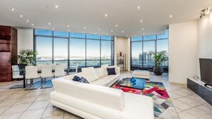 A spacious living room with a large white sofa, a coffee table, and a view of the city through large windows. - Exclusive Stays - Gallery Penthouse (Southbank)