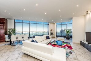 A spacious living room with a large white sofa, a coffee table, and a view of the city through large windows. - Exclusive Stays - Gallery Penthouse (Southbank)