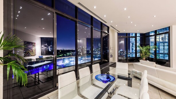 A modern interior with a glass table, white chairs, and a view of a cityscape at night. - Exclusive Stays - Gallery Penthouse (Southbank)