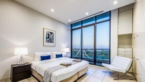 A modern bedroom with a large bed, a nightstand, a chair, and a view of a bridge through large windows. - Exclusive Stays - Gallery Penthouse (Southbank)