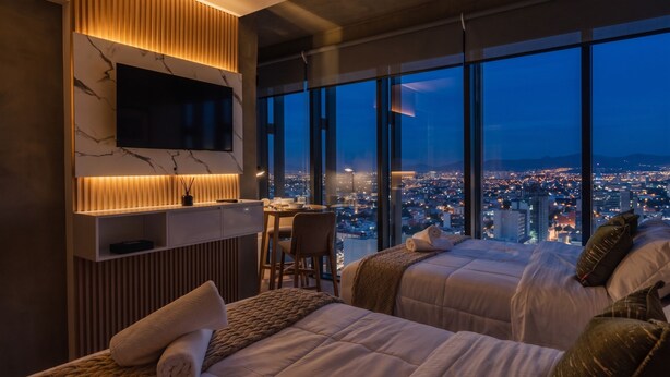 Ein modernes Hotelzimmer mit großem Fernseher, zwei Betten, einem Schreibtisch und Blick auf die Stadt.