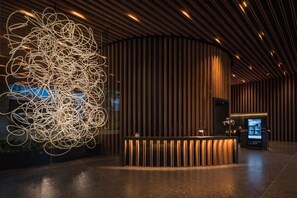 Una zona de recepción moderna con un mostrador curvo de madera, una gran instalación de luces abstractas y un quiosco informativo digital.