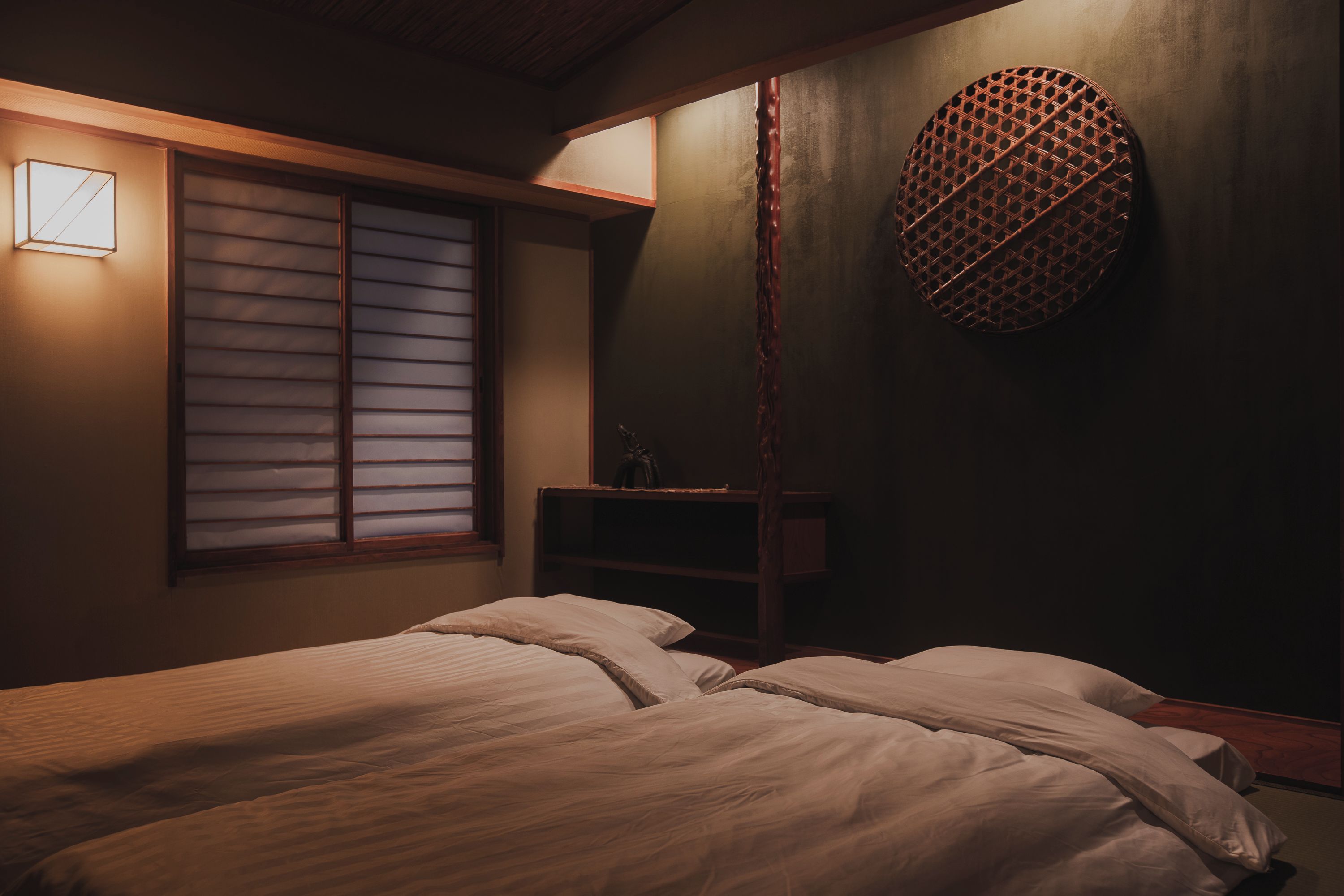Une chambre à coucher avec un lit, une fenêtre avec des stores, un objet circulaire fixé au mur et une étagère sur laquelle se trouve un petit objet.