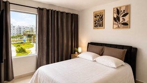 A hotel room with a large window, a bed with white linens, a nightstand with a lamp, and two wall art pieces. - Apto moderno en Serena con balcon  (Cartagena)