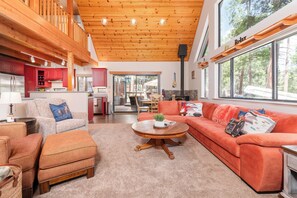 A spacious living area with a fireplace, a kitchen with red cabinets, and large windows offering a view of the outdoors. - Cozy Mountain Retreat, 5 Min Walk to  Downtown, Lake Membership, Sleeps 7 (Twain Harte)