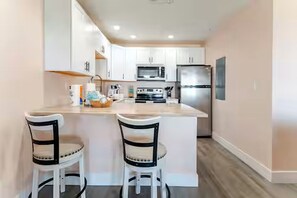 A modern kitchen with white cabinets, a central island with stools, a microwave, stove, and refrigerator. - Adorable 1-bedroom condo steps from the beach in Perdido Key - Sleeps 6!! (Pensacola)