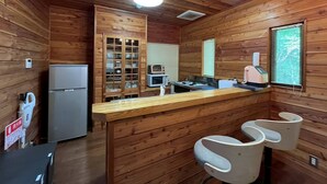 Interior de una cabaña de madera con kitchenette, refrigerador, microondas, estufa y una ventana con persianas.