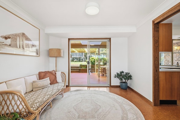 Habitación con sofá de mimbre, alfombra estampada, planta en maceta y vista a un patio exterior con mesa y sillas.