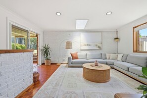 Una sala de estar moderna con un sofá seccional gris, una mesa de centro de mimbre redonda, una pared de ladrillo blanco y un tragaluz.