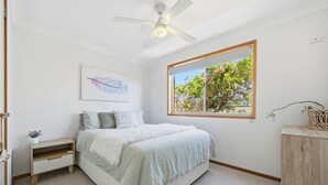 A bedroom with a bed, a ceiling fan, a window with a view of trees, a wall-mounted artwork, and a small bedside table.