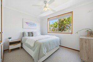 Un dormitorio con cama, ventilador de techo, ventana con vistas a árboles, una obra de arte en la pared y una mesita de noche pequeña.