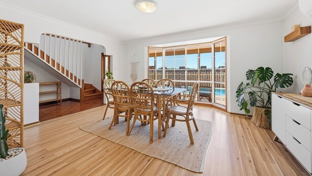 A spacious room with a wooden floor, a dining area with wicker chairs, a staircase, and a large window with a view of a pool.