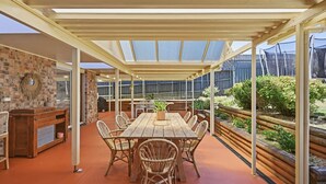 A covered patio with a wooden table and wicker chairs, a brick wall with a decorative wreath, and a garden area with plants and a wooden fence.
