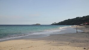 Una playa con orilla arenosa, una pequeña isla a lo lejos y algunas personas caminando.