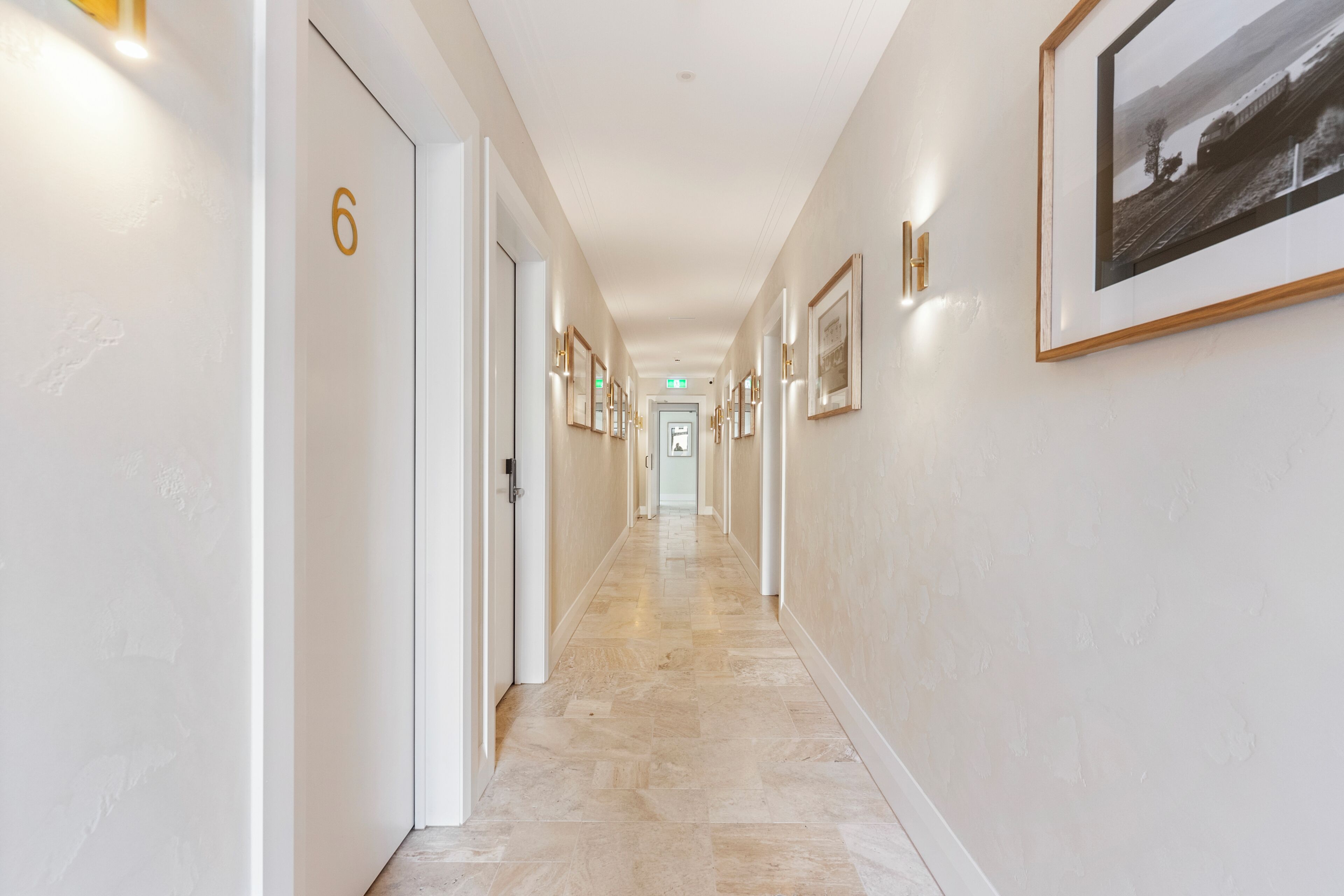 A hotel hallway with a marble floor, numbered doors, framed pictures on the walls, and a framed photograph on the right.