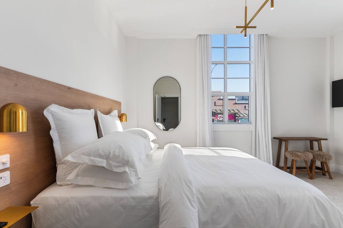 Habitación de hotel con una cama grande, cabecera de madera, mesita de noche con lámpara, una pequeña mesa con una silla y un televisor montado en la pared.