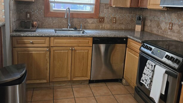 A kitchen with wooden cabinets, a stainless steel dishwasher, a stove, and a window with blinds.